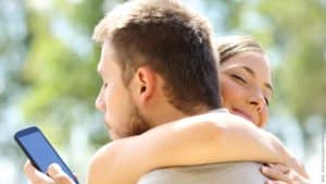 Gefahr für die Beziehung durch das Internet. Mann und Frau umarmen sich. Er schaut hinter dem Rücken der Frau heimlich in sein Handy.