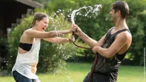 Ist der Mensch überhaupt für eine Partnerschaft gebaut? Junges Paar kämpft um den Gartenschlauch und spritzt sich dabei komplett nass. Beide lachen.