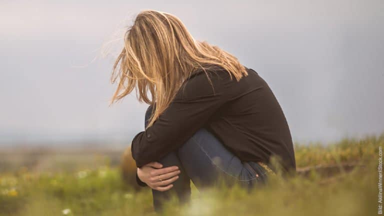 Vergiftete Beziehung: 7 Warnzeichen. Frau sitzt auf einer Wiese in der Hocke. Sie hat ihre Arme um ihre Knie geschlungen und den Kopf gesenkt. Sie wirkt traurig.