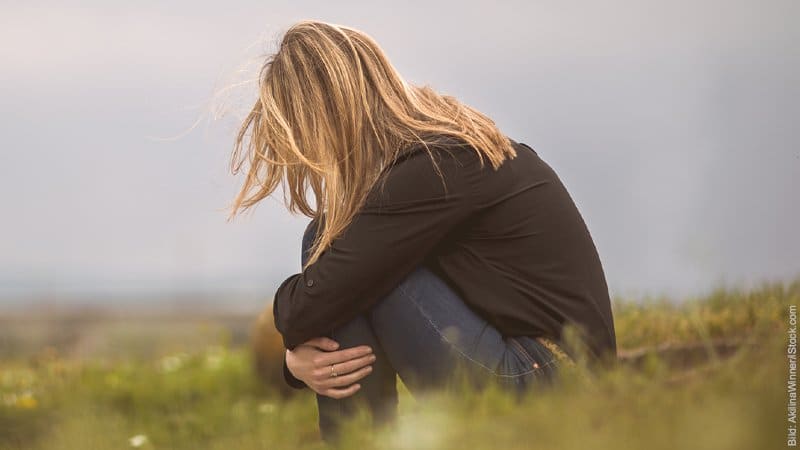 Vergiftete Beziehung: 7 Warnzeichen. Frau sitzt auf einer Wiese in der Hocke. Sie hat ihre Arme um ihre Knie geschlungen und den Kopf gesenkt. Sie wirkt traurig.