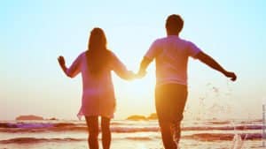 Warum Sie den richtigen Partner nicht finden. Ein Paar hält sich an den Händen und läuft in Richtung Sonnenuntergang am Strand.