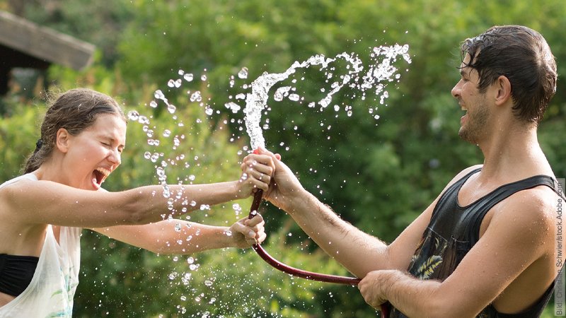 Distanz in Beziehungen – Der Distanz-Pol. Junges Paar neckt sich mit dem Gartenschlauch. Dabei spritze das Wasser um sie herum.