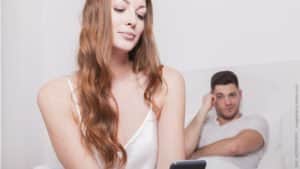 Kann ein Fremdgeher treu werden: 1x Affäre, wieder Affäre? Mann und Frau im Bett; der Mann schaut skeptisch zu seiner Frau, die ins Handy lächelt.