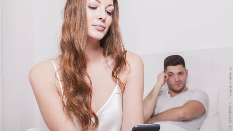 Kann ein Fremdgeher treu werden: 1x Affäre, wieder Affäre? Mann und Frau im Bett; der Mann schaut skeptisch zu seiner Frau, die ins Handy lächelt.
