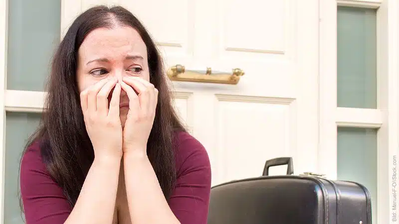 Trennung vorbereiten. Frau sitzt neben Koffer vor dem Haus. Sie hat die Hände vor dem Gesicht und sieht traurig aus.