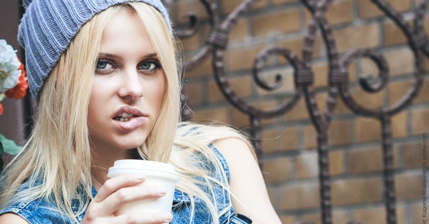 Unterbrechen schwer gemacht – 7 Tipps. Junge blonde Frau mit Kaffee-Becher in der Hand beißt sich auf die Lippe. Sie überlegt offenbar, was sie tun will, wenn sie wieder unterbrochen wird.