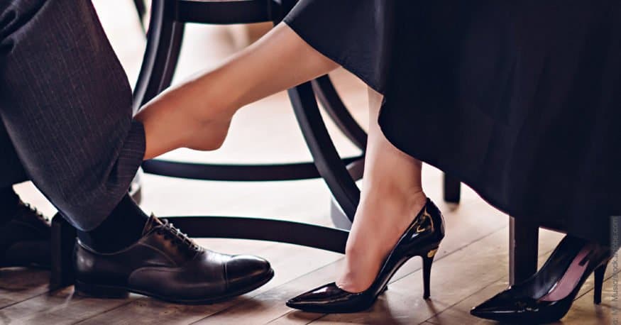 Sexualisierung in der Partnerschaft mal anders herum: Frau hat Schuh ausgezogen und berührt damit einen Mann am Bein unter einem Tisch.