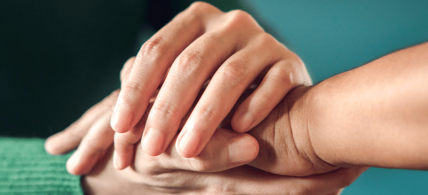 Bild: Eine Hand legt sich über die Hände eines anderen Menschen. Petrolfarbener Hintergrund. Vertrauen nach dem Fremdgehen – so funktioniert Heilung wirklich