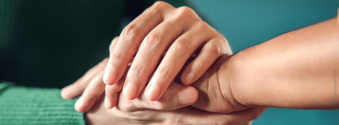 Bild: Eine Hand legt sich über die Hände eines anderen Menschen. Petrolfarbener Hintergrund. Vertrauen nach dem Fremdgehen – so funktioniert Heilung wirklich