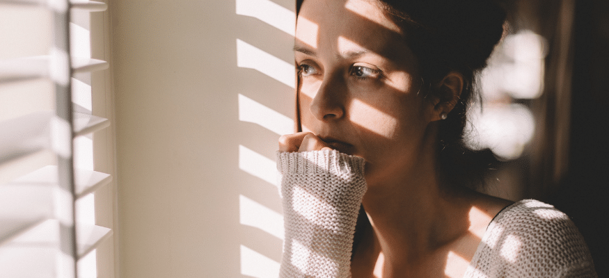 Frau steht sehnsüchtig am Fenster, Schatten der Jalousie auf ihrem Gesicht: Wenn der Gedanke an ihn (oder sie) nicht verschwindet Was tun nach dem Betrug