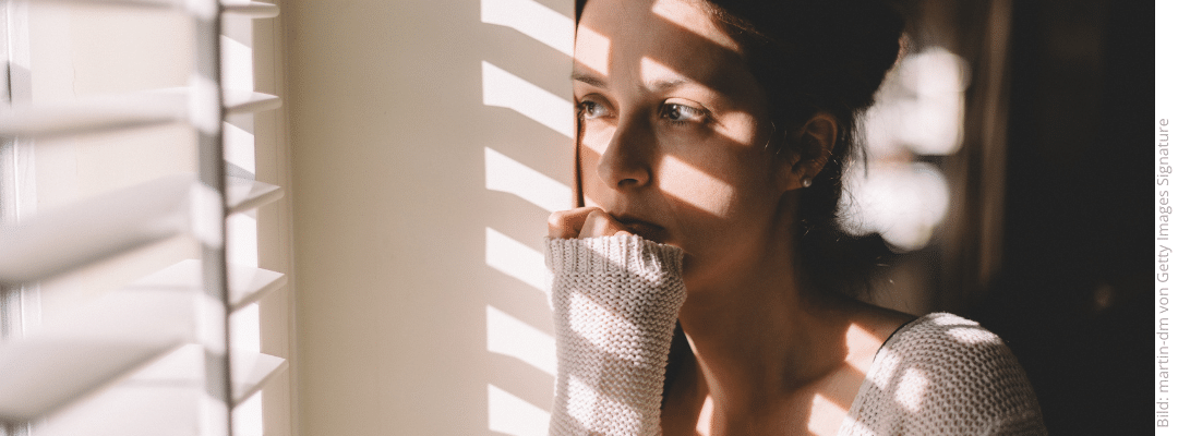 Frau steht sehnsüchtig am Fenster, Schatten der Jalousie auf ihrem Gesicht: Wenn der Gedanke an ihn (oder sie) nicht verschwindet Was tun nach dem Betrug